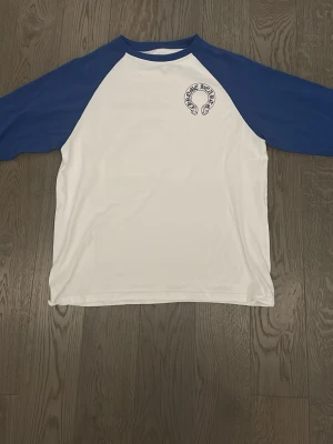 Blåvit Chrome Hearts långärmad t-shirt - Säljer en blåvit t-shirt från Chrome Hearts med blå raglanärmar och tryck både fram och bak. Framsidan har en liten logga på bröstet och baksidan har stora bokstäver samt ytterligare ett tryck nedtill. Fick den i julklapp och säljer eftersom det inte är min stil.