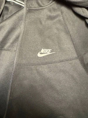 Svart hoodie från Nike med logga - Svart hoodie från Nike med klassisk vit logga på bröstet. Hoodien har huva och långa ärmar, tillverkad i mjukt material som känns skönt mot huden. Perfekt för en avslappnad och sportig stil.