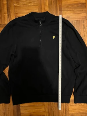 Svart stickad tröja från Lyle & Scott - Svart stickad tröja från Lyle & Scott med halv dragkedja och gul logga på bröstet. Tröjan har ribbade muddar vid ärmslut och nederkant samt en klassisk rak passform. Perfekt till höst och vinter för en stilren look.