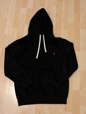 Svart hoodie från Polo Ralph Lauren - Svart hoodie från Polo Ralph Lauren med vit dragsko i huvan och den klassiska röda loggan broderad på bröstet. Hoodien har en avslappnad passform och är tillverkad i mjukt material, perfekt för en chill och stilren look.