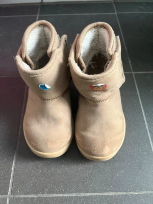 Beige fodrade barnstövlar - Säljer ett par beige barnstövlar med mjukt vitt foder och kardborreknäppning på sidan. Stövlarna har rund tå och dekorationer framtill med djurmotiv. Perfekta för kalla dagar och riktigt sköna att ha på sig.