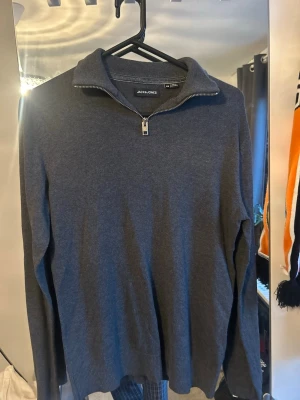 Mörkgrå halvzip tröja Jack & Jones - Mörkgrå långärmad tröja från Jack & Jones med halvzip och hög krage. Tröjan är i mjukt material och har ribbade muddar vid ärmslut och nederkant. Perfekt för lager på lager och enkel att matcha med jeans eller chinos.