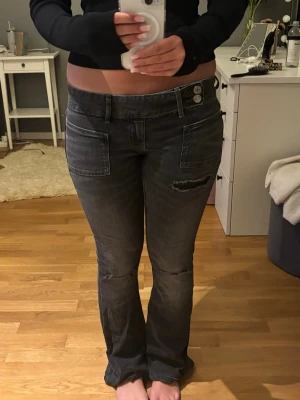 Svarta bootcut jeans med dubbla knappar - Säljer ett par svarta bootcut jeans med dubbla knappar i midjan och snygga fickdetaljer framtill. Jeansen har en klassisk femficksdesign och är tillverkade i ett stretchigt denim-material som sitter bekvämt. Perfekta för dig som gillar en cool och avslappnad stil.