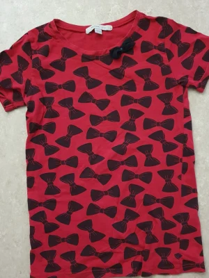 T-shirt - Snygg röd t-shirt från mywear young med svarta rosetter över hela tröjan och en extra tygrosett vid halsringningen. T-shirten har rund hals och korta ärmar. Perfekt för dig som gillar lekfulla mönster och vill sticka ut lite extra.