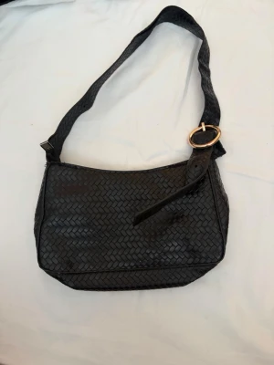 Svart flätad axelväska med spänne - Snygg svart handväska i flätad design med bred axelrem och ett guldfärgat runt spänne. Väskan har en mjuk, rektangulär form och är tillverkad i konstläder. Perfekt för dig som gillar trendiga accessoarer med lite edge.