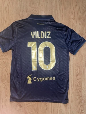 Juventus matchtröja Yildiz #10 - Snygg mörkblå Juventus matchtröja från Adidas med guldiga detaljer och tryck. På ryggen står Yildiz och nummer 10 i stort guldtryck. Framtill finns Save the Children-logga och klubbmärke. Tröjan har krage och korta ärmar, perfekt för fotbollsträning eller match.