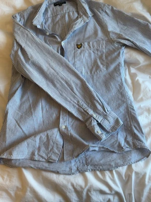Randig skjorta från Lyle & Scott - Snygg blåvit randig skjorta från Lyle & Scott med klassisk krage och knappstängning. Skjortan har lång ärm och en bröstficka med den ikoniska gula loggan. Tillverkad i mjuk bomull för en bekväm känsla.