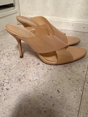 Beige klackade sandaler från Zara - Säljer ett par snygga beige sandaler från Zara med hög klack och breda korslagda remmar i konstläder. Öppen tå och slip-in modell som ger en trendig look. Perfekta för dig som vill ha stilrena och moderna skor till sommaren.