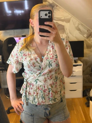 Blommig omlottblus med knytband - Säljer en söt blommig omlottblus i vitt med rosa och gröna detaljer. Blusen har korta ärmar, volangkant och knytband i midjan för en feminin look. Perfekt till jeans eller kjol för en fräsch stil.