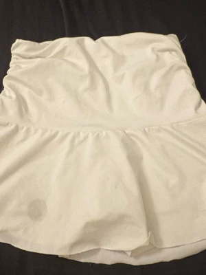 Vit skort med volang - Säljer en vit skort med volangkant och inbyggda shorts under kjoldelen. Skorten har en enkel och stilren design med hög midja och mjukt, stretchigt material. Perfekt för dig som vill ha kjolkänsla men ändå känna dig bekväm. Har en fläck fram, se bild 1, har inte försökt få bort den, därav priset.