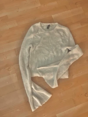 Vit fluffig stickad tröja H&M - Supersöt vit stickad tröja från H&M med fluffig känsla och vida trumpetärmar. Tröjan har rund halsringning och är gjord i ett mjukt, lurvigt material som känns riktigt cozy. Perfekt för dig som gillar en trendig och mysig look.