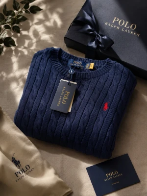 Mörkblå kabelstickad tröja Polo Ralph Lauren - Kabelstickad mörkblå tröja från Polo Ralph Lauren med rund halsringning och röd broderad logotyp på bröstet. Tröjan har ribbstickade muddar och är tillverkad i mjukt material som ger en klassisk och stilren look.
