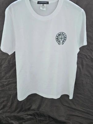 Vit Chrome Hearts t-shirt med tryck - Vit t-shirt från Chrome Hearts med svart tryck på bröstet och stort tryck på ryggen. Klassisk passform och rund halsringning. Perfekt för dig som gillar streetwear och vill sticka ut med en ikonisk design.