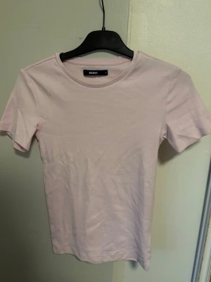 Ljusrosa t-shirt från BikBok - En enkel och stilren ljusrosa t-shirt från BikBok. T-shirten har rund halsringning och korta ärmar. Tillverkad i mjuk bomull som känns skön mot huden. Perfekt basplagg att matcha med allt.