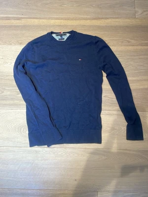 Mörkblå stickad tröja Tommy Hilfiger - Mörkblå stickad tröja från Tommy Hilfiger i bomull. Klassisk rund halsringning och diskret logga broderad på bröstet. Tröjan har ribbade muddar vid ärmslut och nederkant, perfekt för en stilren och enkel look.
