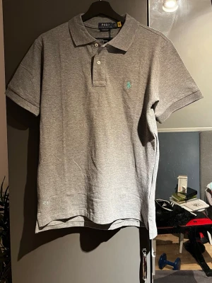 Grå pikétröja Polo Ralph Lauren - Klassisk grå pikétröja från Polo Ralph Lauren med korta ärmar och krage. Tröjan har två knappar vid halsen och den ikoniska broderade loggan i ljusblått på bröstet. Tillverkad i mjuk bomull för en bekväm känsla.