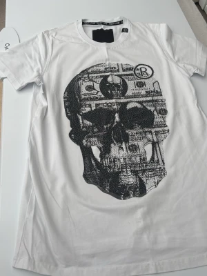 Vit t-shirt med döskalletryck - Cool vit t-shirt med stort svart döskalletryck framtill. T-shirten har rund halsringning och korta ärmar. Perfekt för dig som gillar edgy och unik stil. Storleken är L men är som M