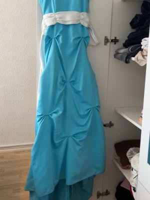 Lång blå klänning med vit knytband - Elegant långklänning i ljusblått med vit knytband i midjan. Klänningen har draperade detaljer och en vacker fallande form. Perfekt för speciella tillfällen där du vill sticka ut med en unik look. Man kan också skjustera längden där nere.