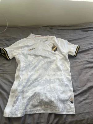 Vit AIK matchtröja från Nike - Säljer en vit AIK matchtröja från Nike med gulddetaljer och klubbmärke på bröstet. Tröjan har ett unikt mönster med Stockholmsmotiv och texten STOCKHOLMENSIS i kragen. Tillverkad i Dri-FIT material som andas, perfekt för träning eller match.