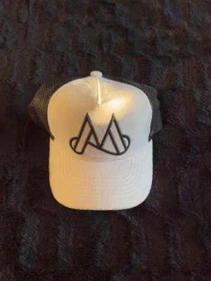 Vit och svart keps med mesh - Säljer en stilren vit keps med svart mesh på sidorna och bak. Framtill finns en stor svart broderad logga med grafiskt mönster. Kepsen har böjd skärm och justerbar snapback. Perfekt för dig som gillar streetwear och enkel design.