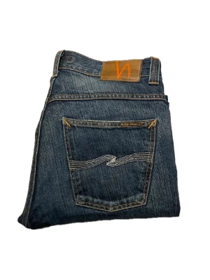 Mörkblå jeans från Nudie Jeans - Snygga mörkblå jeans från Nudie Jeans med klassisk femficksmodell och kontrastsömmar. Bakfickan har den ikoniska vågiga sömmen och läderpatch med logga i midjan. Tillverkade i slitstark denim, perfekta för en avslappnad och trendig look.