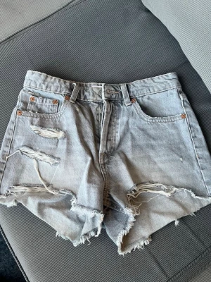 Gråa högmidjade jeansshorts - Säljer ett par gråa högmidjade jeansshorts med slitna detaljer och råa kanter från Zara.. Shortsen har klassisk femficksdesign och knäppning med knappar Perfekta för sommaren och ger en avslappnad vibe.