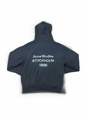 Svart hoodie från Acne Studios - Säljer en svart hoodie från Acne Studios med tryck i vitt på ryggen där det står 'Acne Studios STOCKHOLM 1996'. Hoodien har en klassisk passform med huva och ribbade muddar vid ärmslut och nederkant. Perfekt för en clean och stilren look.