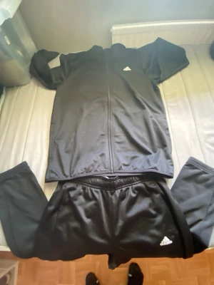 Svart Adidas träningsset i storlek 164! - Säljer ett svart träningsset från Adidas med dragkedja på jackan och vita Adidas-loggor på både jacka och byxor. Setet är gjort i ett lätt och stretchigt material som passar perfekt till träning eller chill. Klassisk sportig look med långärmad överdel och matchande byxor.