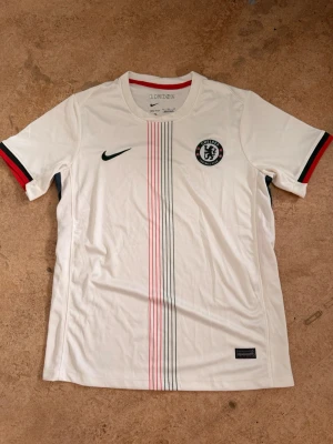 Chelsea vit matchtröja Nike - Chelsea FC matchtröja i vitt med tunna röda och blå ränder längs framsidan. Tröjan har rund hals, korta ärmar med röda och blå detaljer samt Chelsea-logga och Nike Swoosh på bröstet. Tillverkad i lätt och ventilerande material, perfekt för träning eller match.