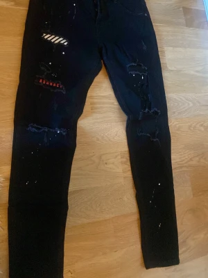 Svarta skinny jeans med slitningar - Säljer ett par svarta skinny jeans med slitningar och patchdetaljer på låret. Jeansen har vita färgstänk och råa hål på båda benen, samt en röd textpatch och en randig patch. Perfekta för en edgy streetstyle-look.