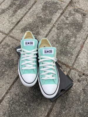 Grönblåa Converse All Star sneakers -  Snygga grönblå/mint/turkos Converse All Star sneakers med klassisk låg modell. Skorna har vit tåhätta i gummi, vita snören och sula samt tydlig All Star logga på plösen. Ovandelen är i canvas och sulan är i gummi. Perfekta för en avslappnad streetstil. De är helt oanvända men ej nyköpta och säljs nu. Original kartongen finns kvar men är lite sliten som bilden visar. 