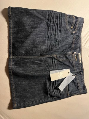 Mörkblå jeanskjol från Denim - Snygg mörkblå jeanskjol från Denim med klassisk femficksdesign och guldfärgade sömmar. Kjolen har en rak passform och är tillverkad i kraftig denim. Perfekt att matcha med en enkel t-shirt eller hoodie.