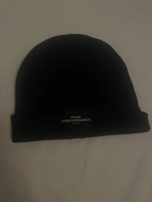 Svart mössa Peak Performance - Snygg svart mössa från Peak Performance med uppvikt kant och diskret logotyp framtill. Klassisk och enkel design som passar till det mesta. Tillverkad i mjukt material som håller dig varm under kalla dagar.