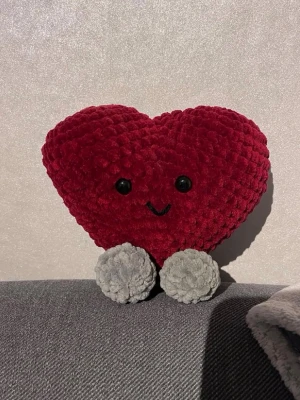 Virkat hjärta❤️ - Söt plyschnalle formad som ett hjärta i mörkröd färg med två stora svarta ögon och ett broderat leende. Hjärtat har två gråa ben och är gjord i mjukt, fluffigt material. Perfekt som dekoration eller gosedjur❤️ den är 13cm på bredden och 16cm på längden🫶🏻