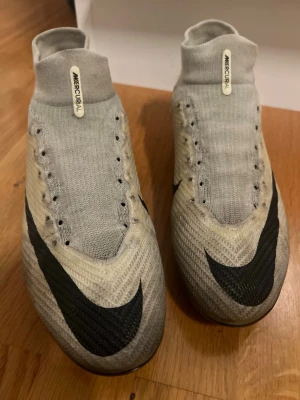 Nike Mercurial vita fotbollsskor - Säljer ett par Nike Mercurial fotbollsskor i vitt med svarta detaljer och hög strumpkrage. Skorna har en modern, strömlinjeformad design och är tillverkade i ett lätt syntetmaterial för bästa bollkontroll och grepp på planen. Perfekta för dig som vill sticka ut på matchen.