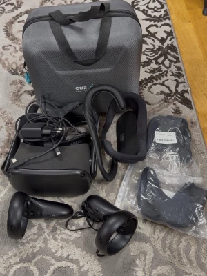 Oculus Quest VR-headset med tillbehör - Säljer ett Oculus Quest VR-headset i svart med två handkontroller, laddare, extra ansiktsfästen och en grå bärväska från CURV. Allt du behöver för att dyka in i VR-världen! Perfekt för gaming och upplevelser i 360 grader.