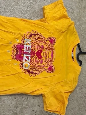 Gul Kenzo t-shirt med tigertryck - Snygg gul t-shirt från Kenzo med ikoniskt tigertryck i rosa och rött framtill samt texten 'KENZO PARIS'. T-shirten har rund halsringning och korta ärmar. Perfekt för dig som vill sticka ut med färg och cool design.