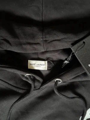 Saint Laurent  - Exklusiv hoodie från Saint Laurent i svart – en tidlös och lyxig piece med den ikoniska loggan framtill. Perfekt för en stilren och modern look med premiumkänsla. 🔺 Storlek S – slim passform ⚜️ Helt nytt skick med tagg 🔺 Tillverkad i mjuk bomull av hög kvalitet 📦 Fraktas inom 24 timmar & trygg affär