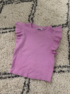 Ljuslila ribbad topp från Zara - Supersöt ljuslila topp från Zara med ribbad struktur och volangdetaljer på axlarna. Toppen är ärmlös och har en rund halsringning. Perfekt för dig som vill ha en feminin och trendig look. (Skriv för fler bilder💕) 