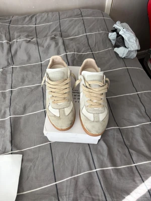Vita och beige sneakers Maison Margiela - Snygga sneakers från Maison Margiela i vitt och beige med klassisk låg siluett. Ovandelen är i en mix av mocka och läder, med beige snörning och gummisula i brunt. Perfekta för dig som gillar stilrena och tidlösa sneakers. Spår av användning finns men det mesta går att tvätta bort.