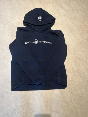 Mörkblå hoodie från Sail Racing - Mörkblå hoodie från Sail Racing med vit logga och text på bröstet. Klassisk huva och magficka framtill. Perfekt för en avslappnad och sportig stil. Tillverkad i mjukt material som känns skönt mot huden.