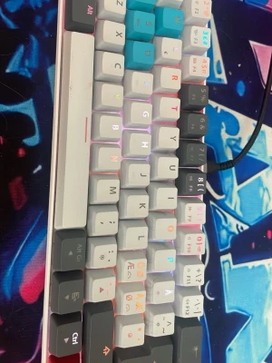 Mekaniskt tangentbord RGB 60% - Säljer ett kompakt mekaniskt tangentbord i 60%-layout med RGB-belysning. Tangenterna är i vitt, grått och turkost med färgglada tecken. Tangentbordet har USB-anslutning och keycaps i plast. Perfekt för gaming eller skrivande på liten yta.