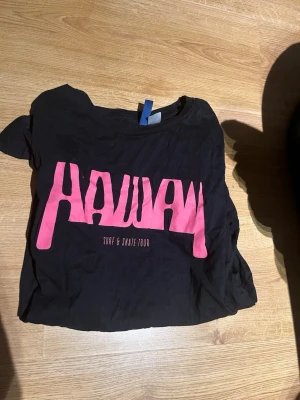 Svart Hawaii t-shirt med tryck - Cool svart t-shirt med stort rosa 'HAWAII' tryck framtill och färgglad palm- och strandgrafik på baksidan. Skön passform och tillverkad i mjuk bomull, perfekt för en avslappnad stil. Textdetaljer som 'Surf & Skate Tour' och platser på Hawaii ger extra vibe.