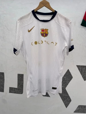 Vit FC Barcelona matchtröja Nike - Snygg vit FC Barcelona matchtröja från Nike med guldfärgade detaljer och klubbmärke på bröstet. Tröjan har mörkblå krage och ärmslut, samt tryck med Lamine Yamal och nummer 10 på ryggen. Tillverkad i lätt och ventilerande material, perfekt för fotbollsträning.