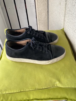 Svarta Lanvin sneakers i mocka - Stilrena svarta sneakers från Lanvin i mocka med matchande svarta skosnören och vit sula. Klassisk låg modell med rund tå och diskret logga på sidan. Perfekta för dig som gillar minimalistisk och tidlös design.