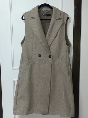 Beige lång väst från ZARA - Stilren beige lång väst från ZARA med dubbelknäppning framtill och breda slag. Västen har två snedställda fickor och är ärmlös med rak passform. Perfekt för lager-på-lager och ger en clean look. Materialet ser ut att vara en mjuk ullblandning.