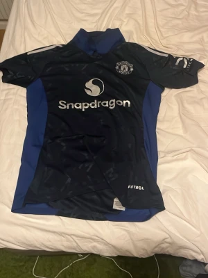 Manchester United bortatröja B. Fernandes - Snygg mörkblå Manchester United bortatröja med B. Fernandes och nummer 8 på ryggen. Tröjan har vita tryck, Snapdragon-logga på bröstet och blå sidopaneler. Perfekt för fotbollsfans som vill sticka ut på planen eller läktaren.