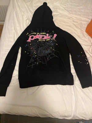 Svart hoodie från Spider med tryck - Svart hoodie från Spider med stor huva och färgglatt tryck på bröstet som inkluderar texten 'around the spiders' och ett spindelnätsmotiv. Ärmarna har små färgglada detaljer och materialet känns mjukt och bekvämt. Perfekt för dig som gillar streetwear och unika prints.