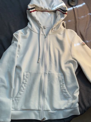 Vit hoodie med dragkedja från Moncler - Snygg vit hoodie från Moncler med dragkedja framtill och två fickor. Huvan har dragsko och insidan är dekorerad med röd och marinblå rand. Moncler-logga på vänster ärm. Tillverkad i mjuk bomullsblandning för skön känsla.