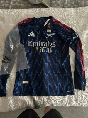 Arsenal långärmad matchtröja blå - Snygg långärmad Arsenal matchtröja från Adidas i mörkblått med blixtmönster, röda ränder på ärmarna och vita detaljer. Tryck med 'Emirates Fly Better' på bröstet och 'GYÖKERES 14' på ryggen. Perfekt för fotbollsfans som vill sticka ut på planen.
